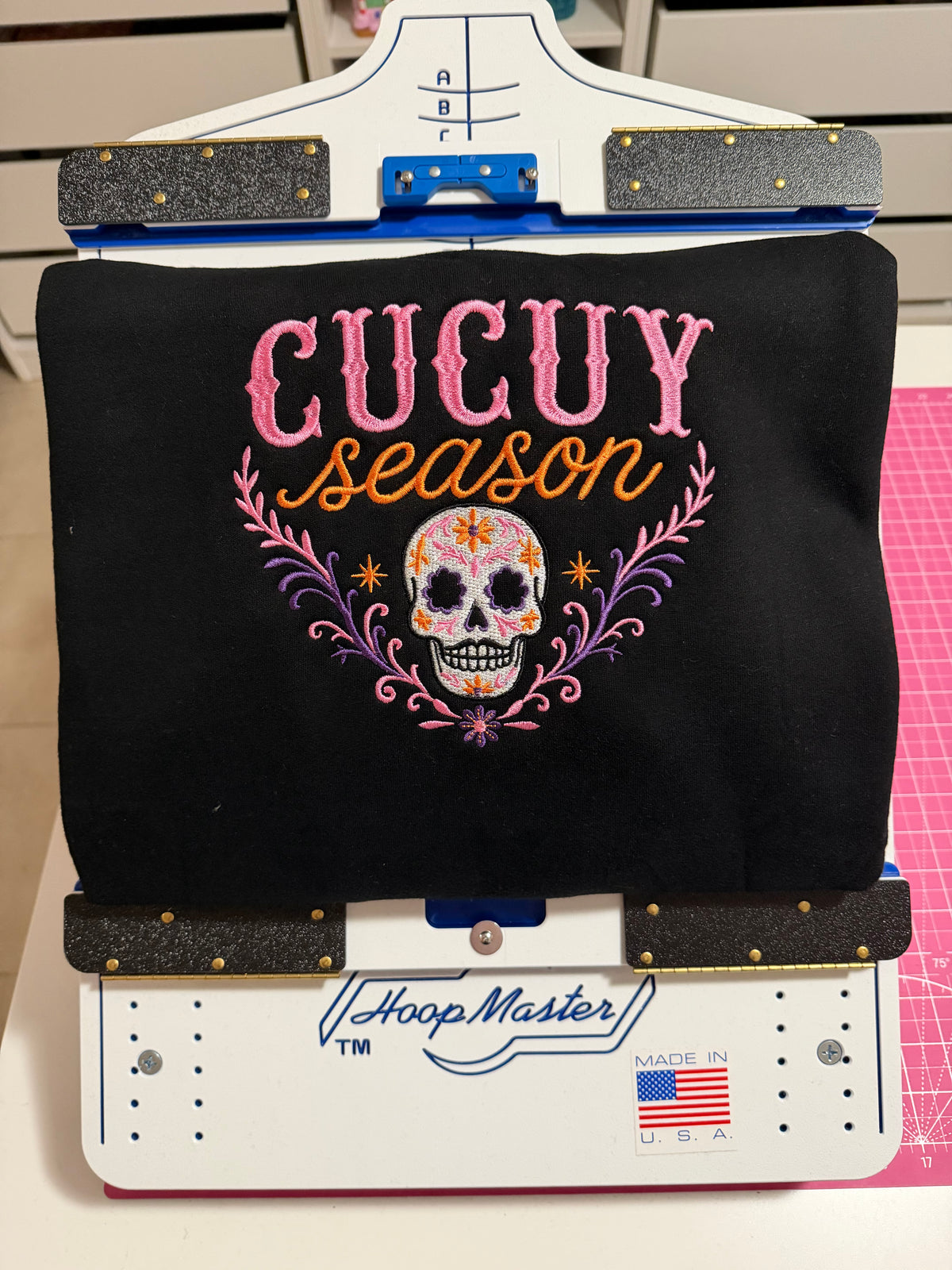 Finished black Cucuy Season hoodie featuring a colorful embroidered sugar skull design displayed on a HoopMaster embroidery station.