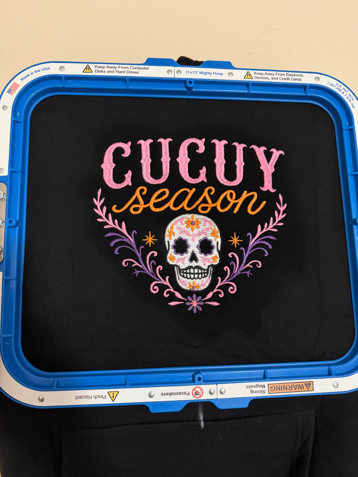 Colorful Cucuy Season sugar skull embroidery stitched on a black hoodie while secured in an embroidery hoop.